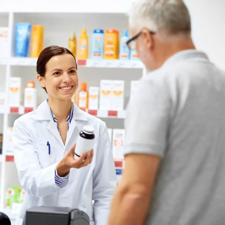 A smiling pharmacist hands a medication bottle to an older patient at the counter