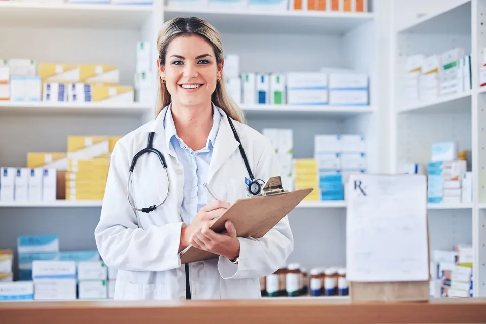 A pharmacist in a white coat holding a clipboard in a pharmacy aisle